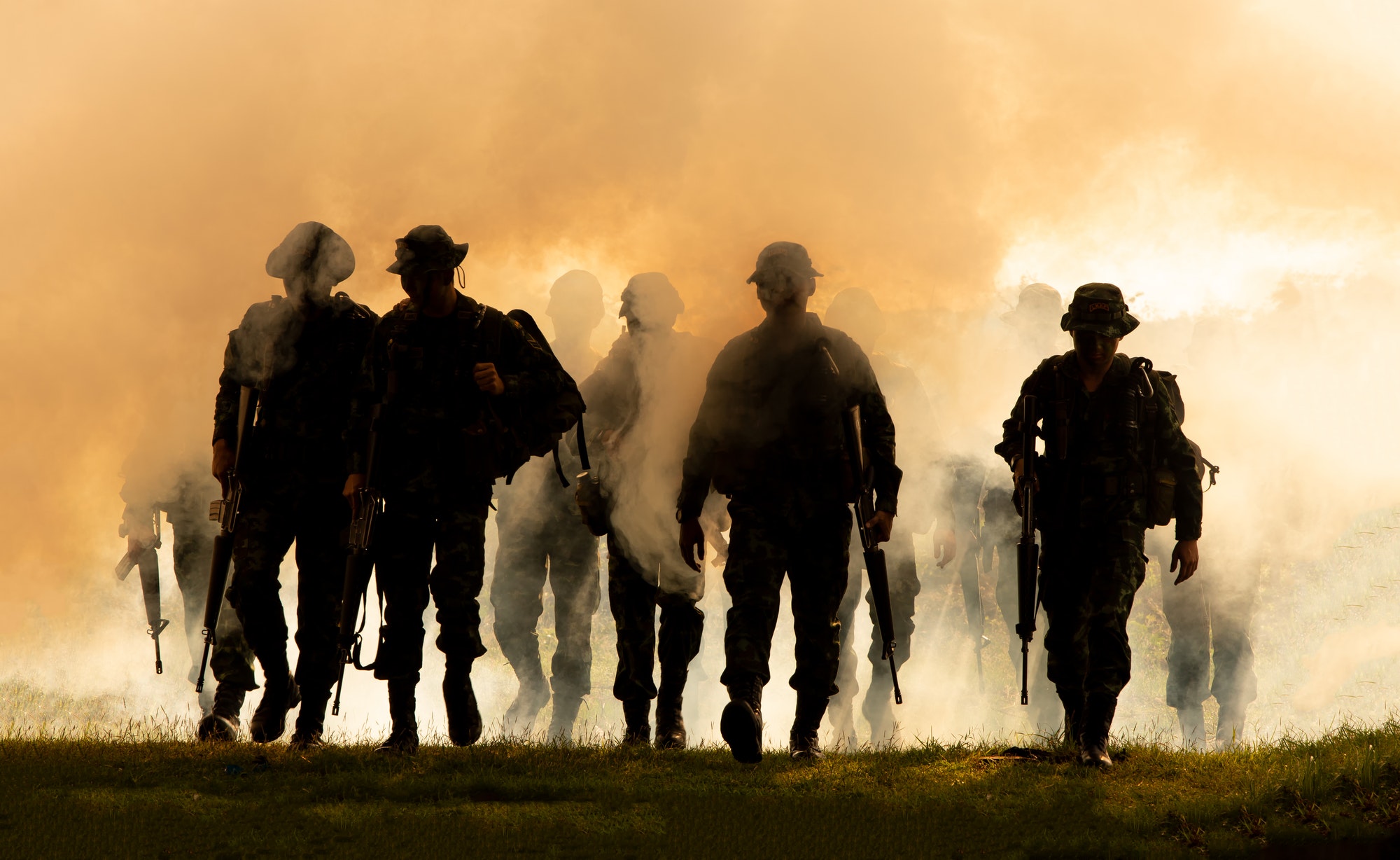 Silhouette of soldiers walking against smoke and sunset background.