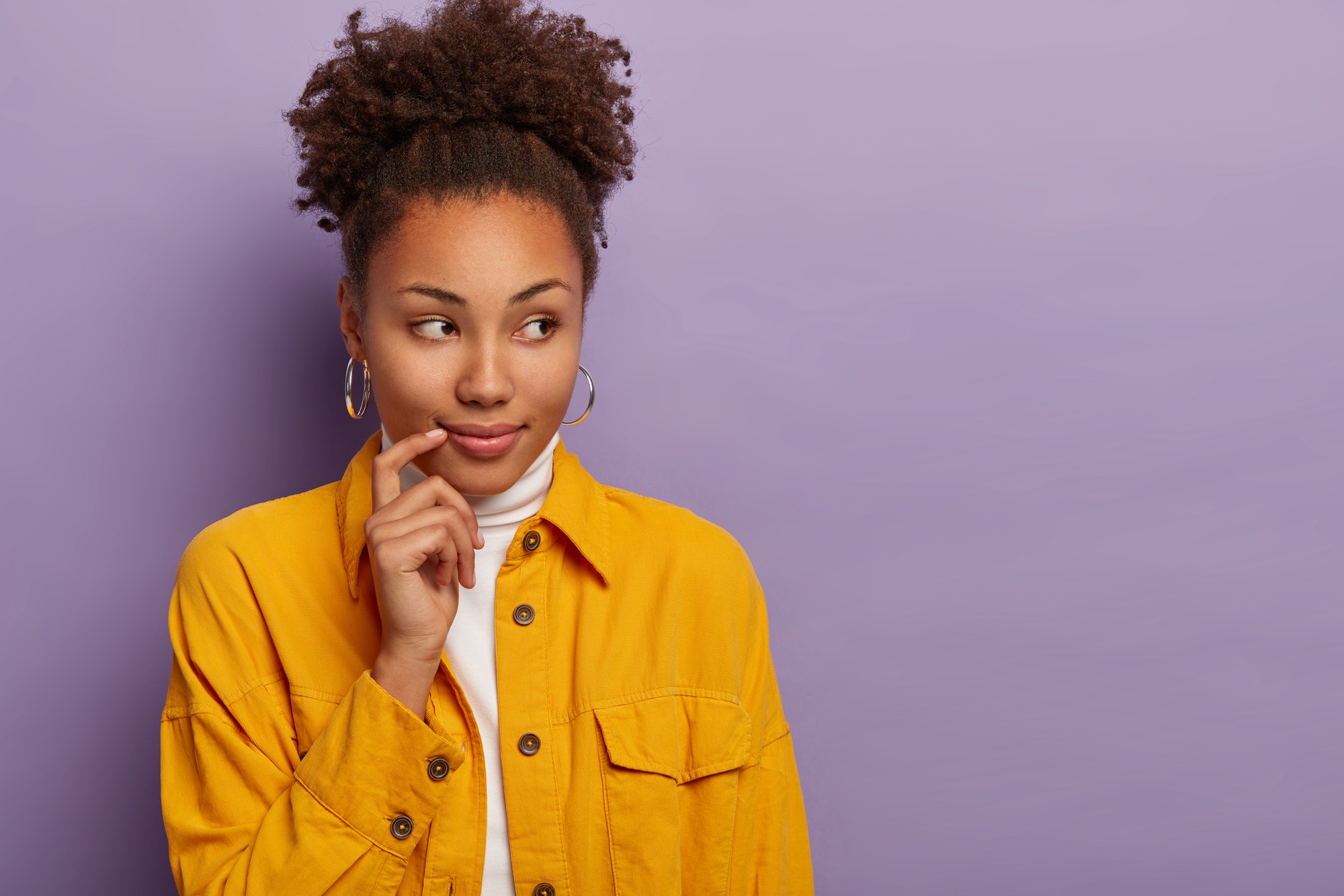 Curly Afro American woman makes decision or assumption, looks aside, wears silver earrings and yello