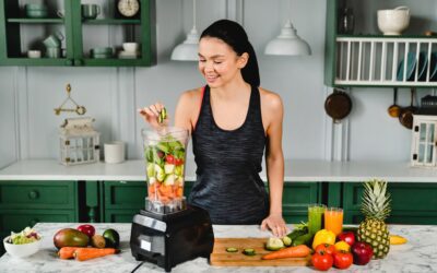 Healthy slim girl adding cucumber to the blender full of fresh vegetables for making healthy