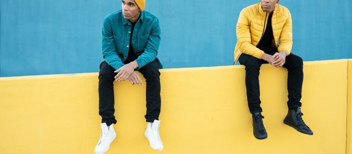 Funny black twin brothers wearing winter clothes, sitting on yellow and blue wall