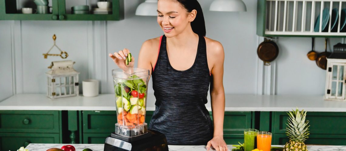 Healthy slim girl adding cucumber to the blender full of fresh vegetables for making healthy