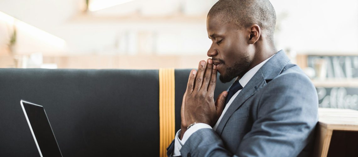 smiling african american businessman with closed eyes praying with laptop