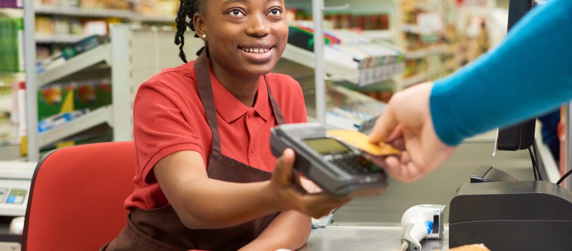 Young smiling salesperson looking at consumer holding credit card over terminal